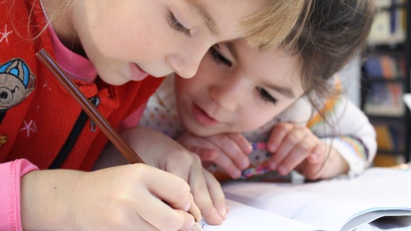 Zwei Grundschulkinder beugen sich über ein Heft. Eines schreibt mit einem Bleistift, das andere schaut aufmerksam zu.