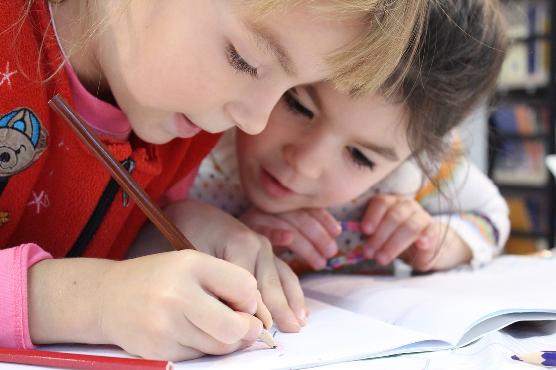 Zwei Grundschulkinder beugen sich über ein Heft. Eines schreibt mit einem Bleistift, das andere schaut aufmerksam zu.