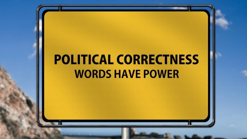 Gelbes Ortseingangsschild mit der Aufschrift „Political Correctness – Words Have Power“ vor einer kurvigen Straße und blauem Himmel.