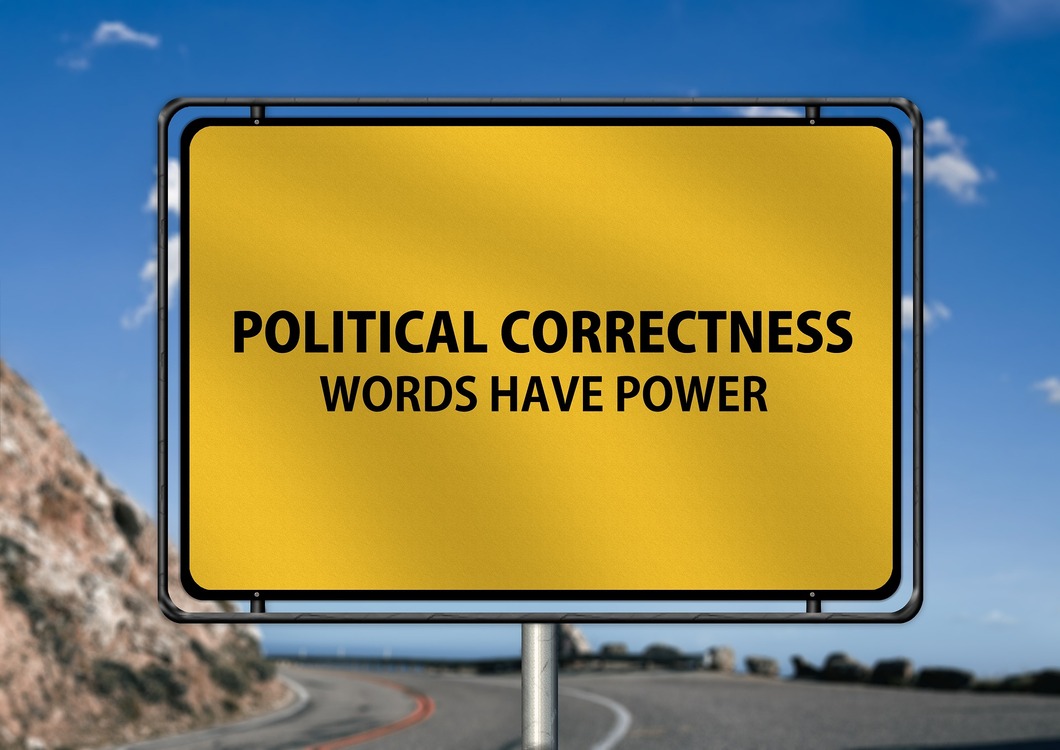Gelbes Ortseingangsschild mit der Aufschrift „Political Correctness – Words Have Power“ vor einer kurvigen Straße und blauem Himmel.